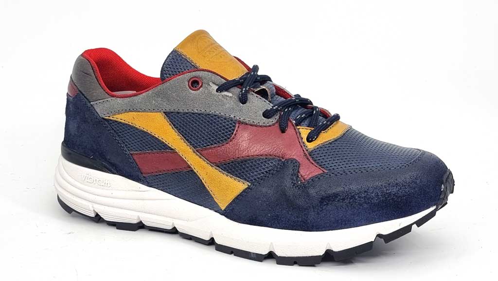 An angled shot displaying the sneaker's side profile and front. The dynamic angle emphasizes the contrast of colors, with yellow and grey details blending seamlessly into the blue leather. The red accent forms a stylish cross near the heel, adding a unique visual element. The slightly elevated rubber sole is visible, providing a glimpse of its flexibility and grip.