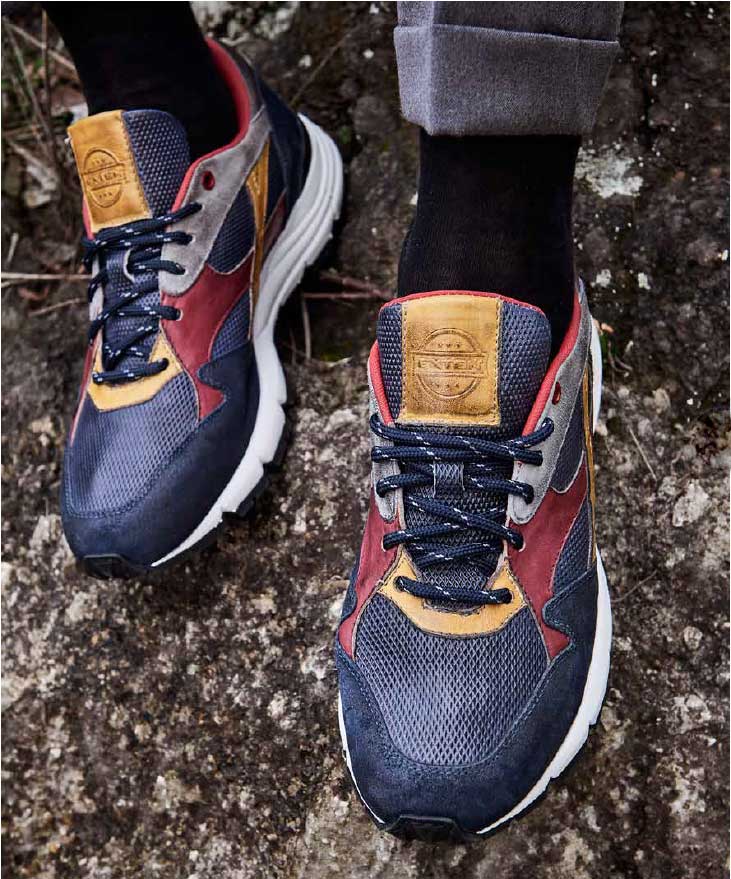 A lifestyle shot of a man wearing the Exton Rocco sneakers on a rugged mountain path. The image captures an adventurous spirit, with the sneakers providing a stylish yet functional choice for outdoor exploration. The durable sole grips the uneven terrain, while the breathable leather upper ensures comfort on the move. The natural lighting enhances the rich blue hue of the sneakers, making them stand out against the earthy landscape.